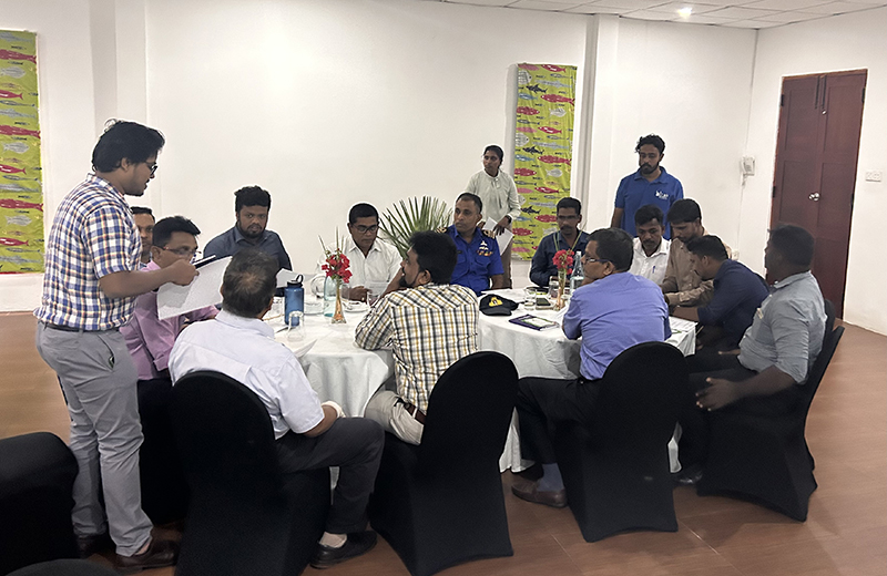 a group of people sat down around a table listening to a person speaking who is standing at the head of the table in a workshop setting