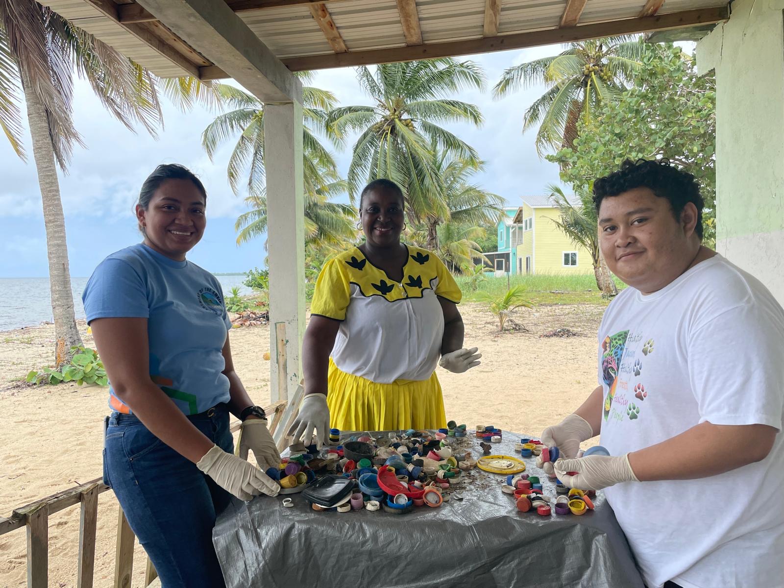 three people standing around a table wearing white gloves, sorting through a range of plastic pieces, all different shapes, colours and sizes, with palm trees and sand in the background