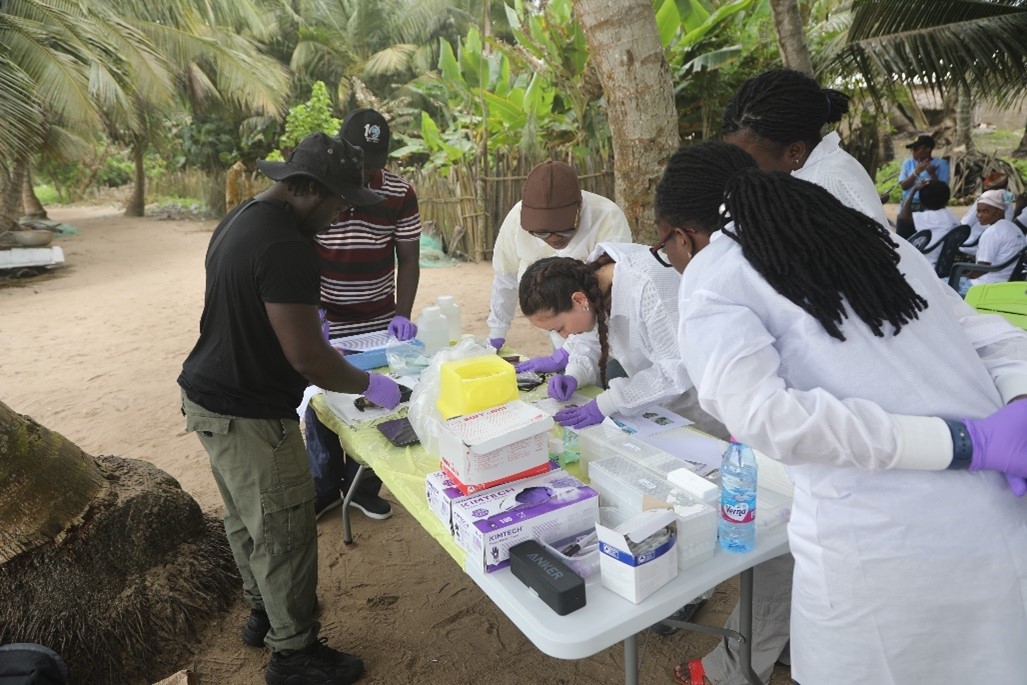 a group of scientists in lab coats standing around a table with various scientific items, watching a scientist demonstrate a technique