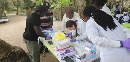 a group of scientists in lab coats standing around a table with various scientific items, watching a scientist demonstrate a technique