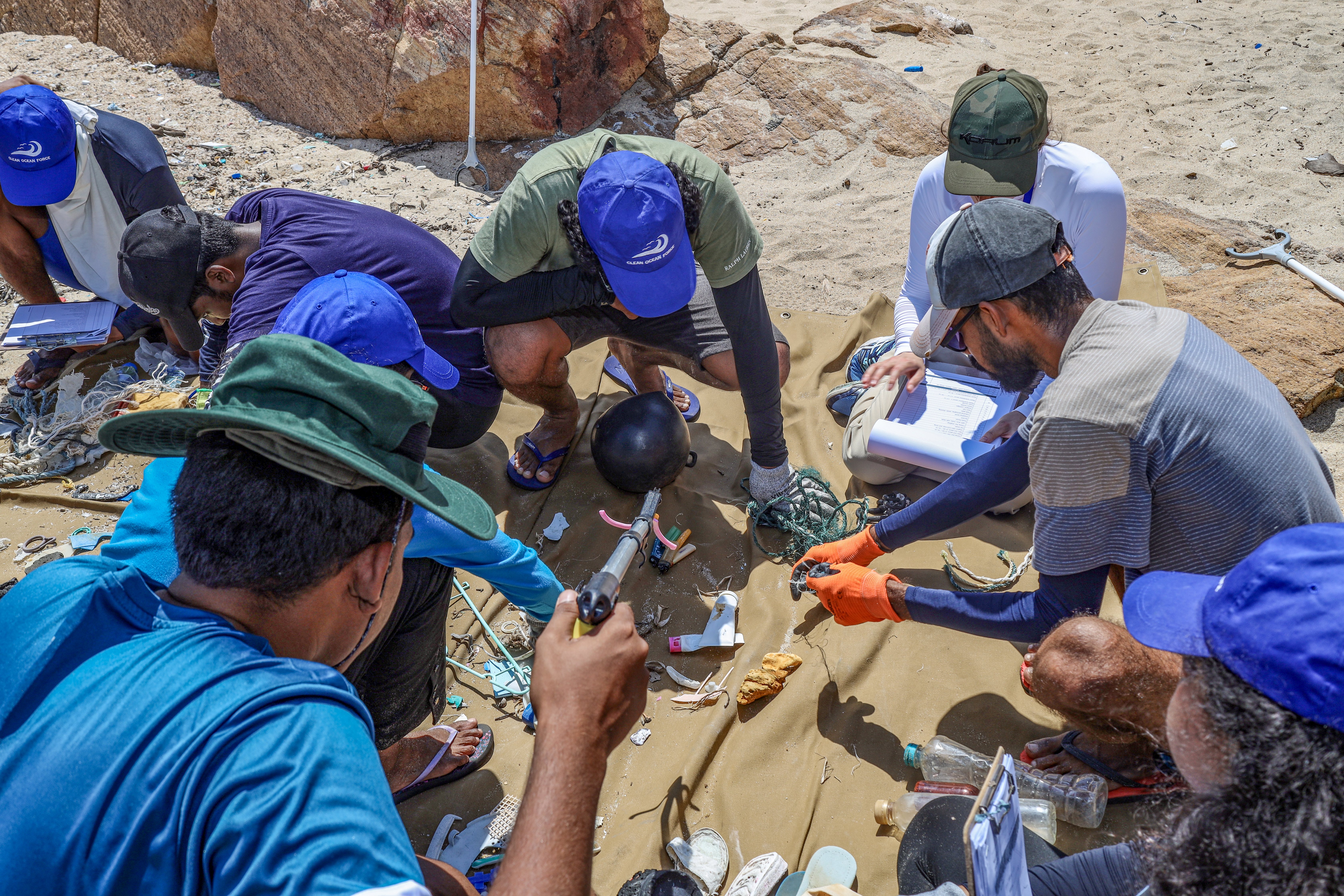 A group of people on a beach gathered around a blanket which has numerous pieces of litter on it, which they have collected.
