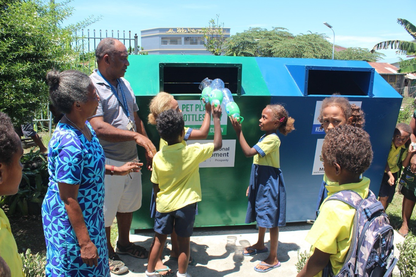 A group of school children standing in front of two large recycling bins with three children putting plastic bottles into the green side. The other side is blue. There are two teachers standing watch.