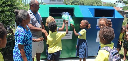 A group of school children standing in front of two large recycling bins with three children putting plastic bottles into the green side. The other side is blue. There are two teachers standing watch.