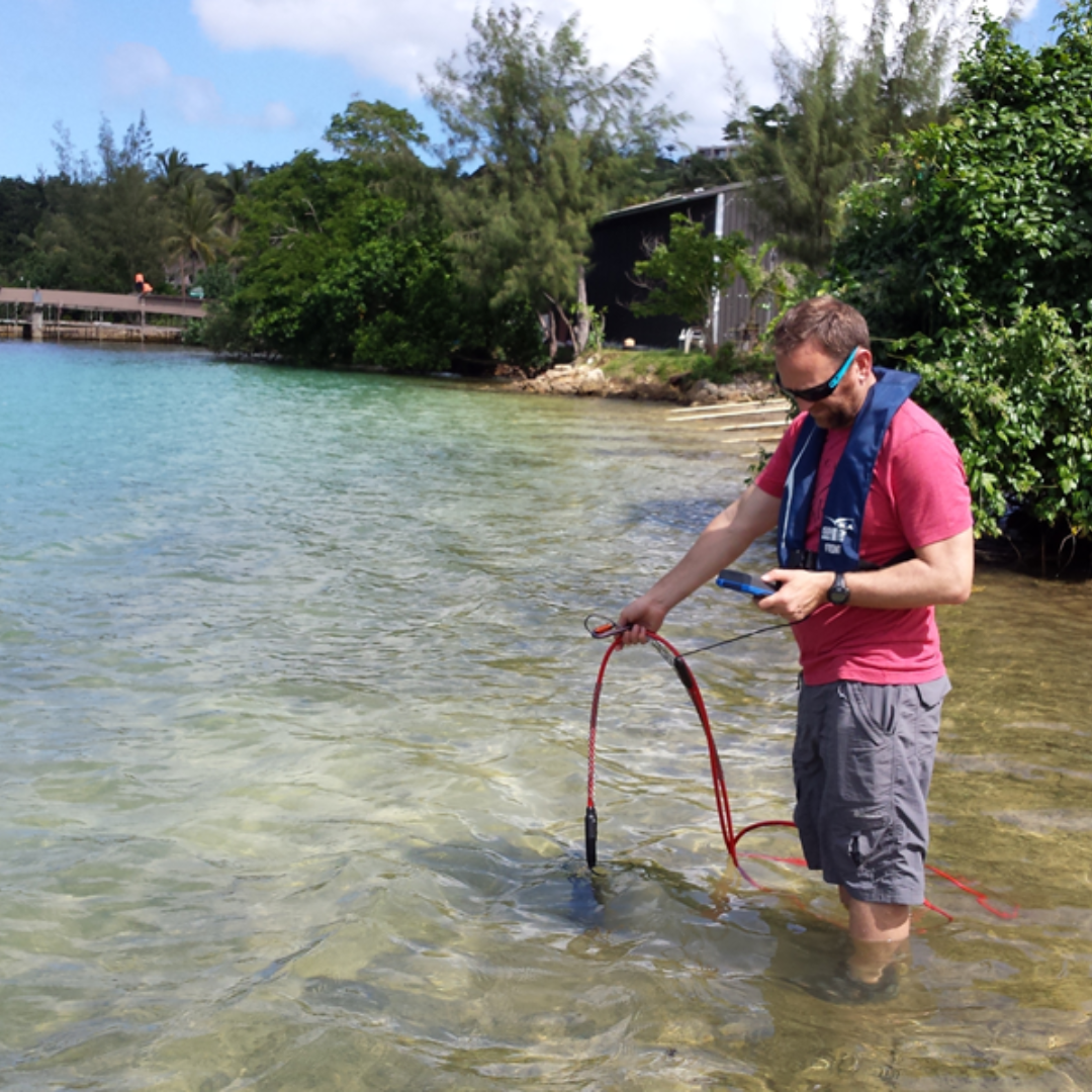 a man standing in shallow water wearing a lifejacket, holding a long probe into the water, and looking at a small screen it is attached to, measuring something
