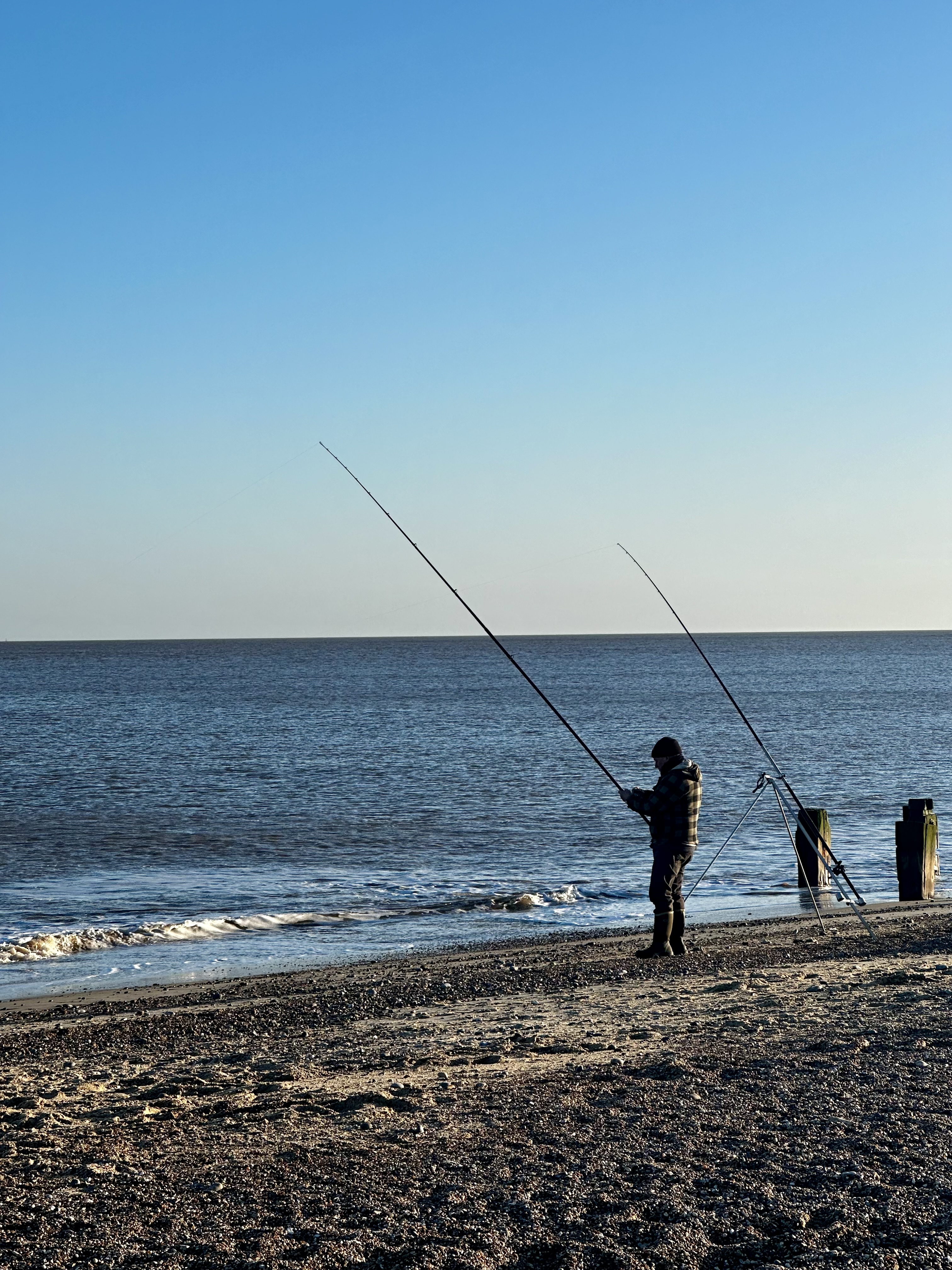 a person standing on a shingle beach holding a fishing rod, with the line in the sea