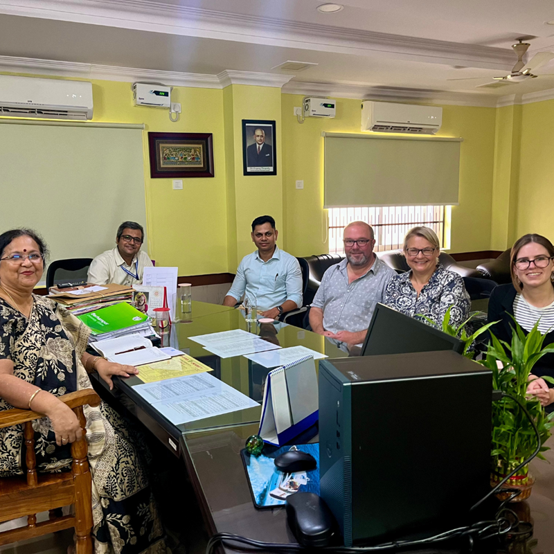 A group of people sat around a table with work books, documents, and laptops, smiling at the camera