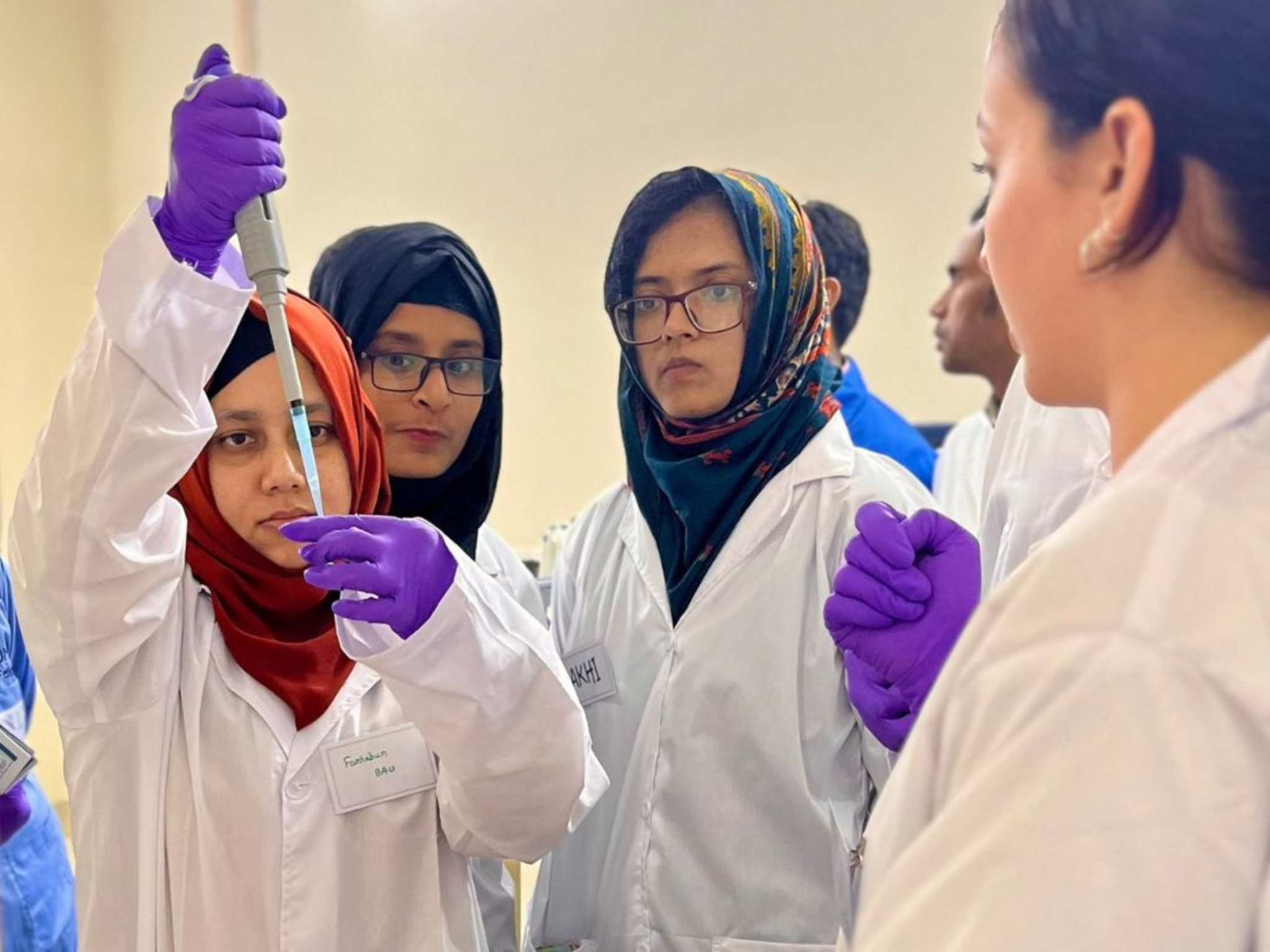 students in white lab coats and purple gloves watching one student with a large pipet conducting an experiment