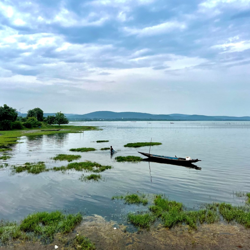 An image of a long boat in water with a person wading in front of it. It is a cloudy day and there is land in the background.
