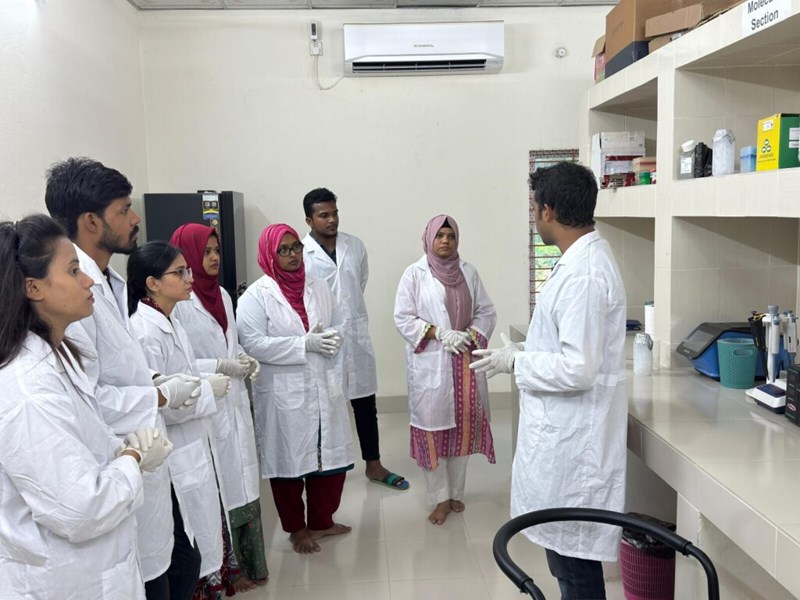 students in white lab coats in a lab setting listening to a teacher who is standing in front of them