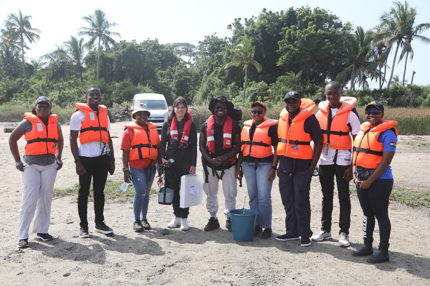 A group of people wearing life jackets standing next to each other and smiling on a beach. One is holding some equipment and there is an industrial bucket on the floor.