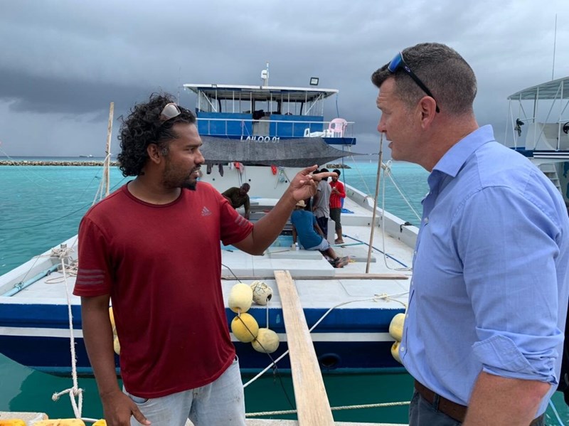 two men are having a conversation in front of a boat which is docked. One of them is pointing behind the other.