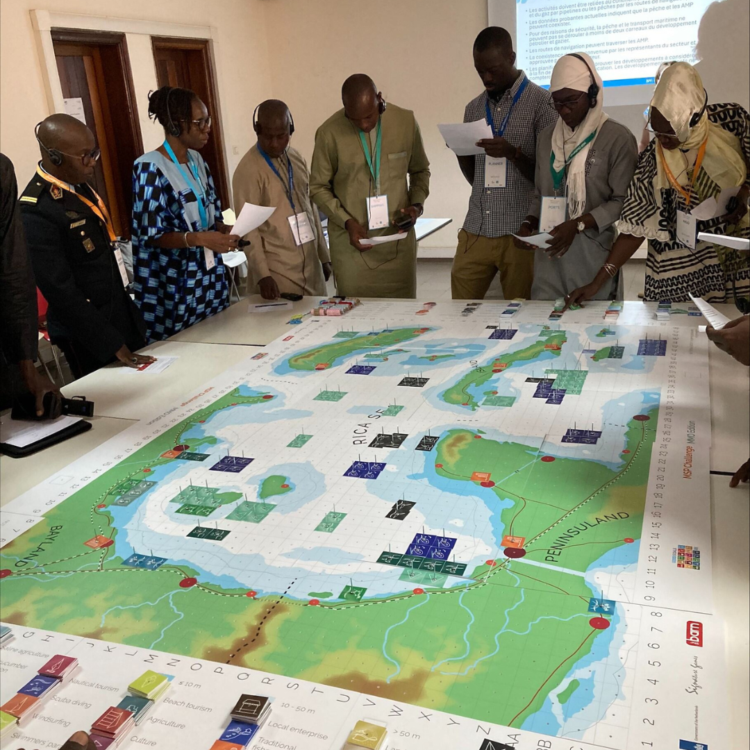 a group of people standing around a large board game, participating in a Marine Spatial Planning exercise