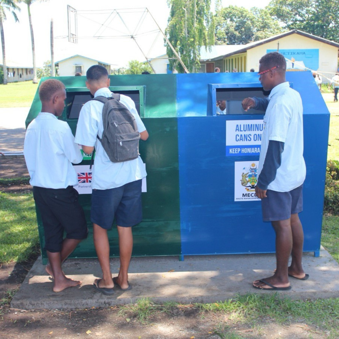 a group of older students putting cans and bottles into a large recycling bin. One side is green for plastic, and the other is blue for aluminum cans