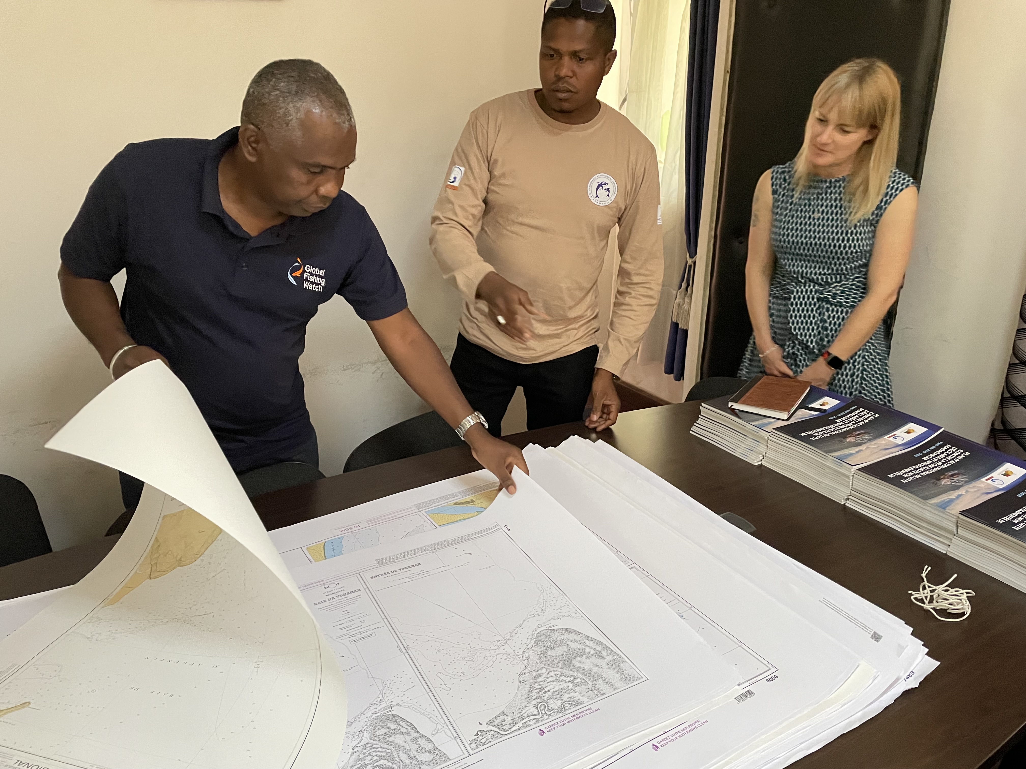 three people standing at a table looking at charts on large pieces of paper, which one person is flicking through