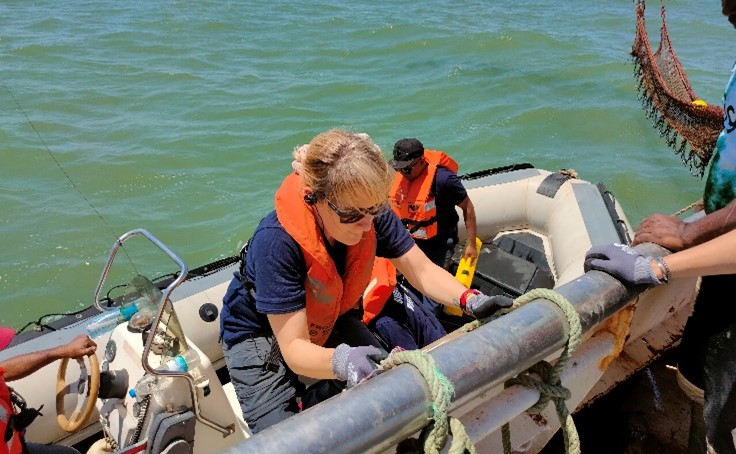 A woman in a uniform and lifejacket is lowering herself down onto a speed boat from a fishing boat. There is a man standing behind her already on the speedy boat, also wearing a lifejacket. 