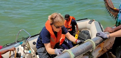 A woman in a uniform and lifejacket is lowering herself down onto a speed boat from a fishing boat. There is a man standing behind her already on the speedy boat, also wearing a lifejacket.