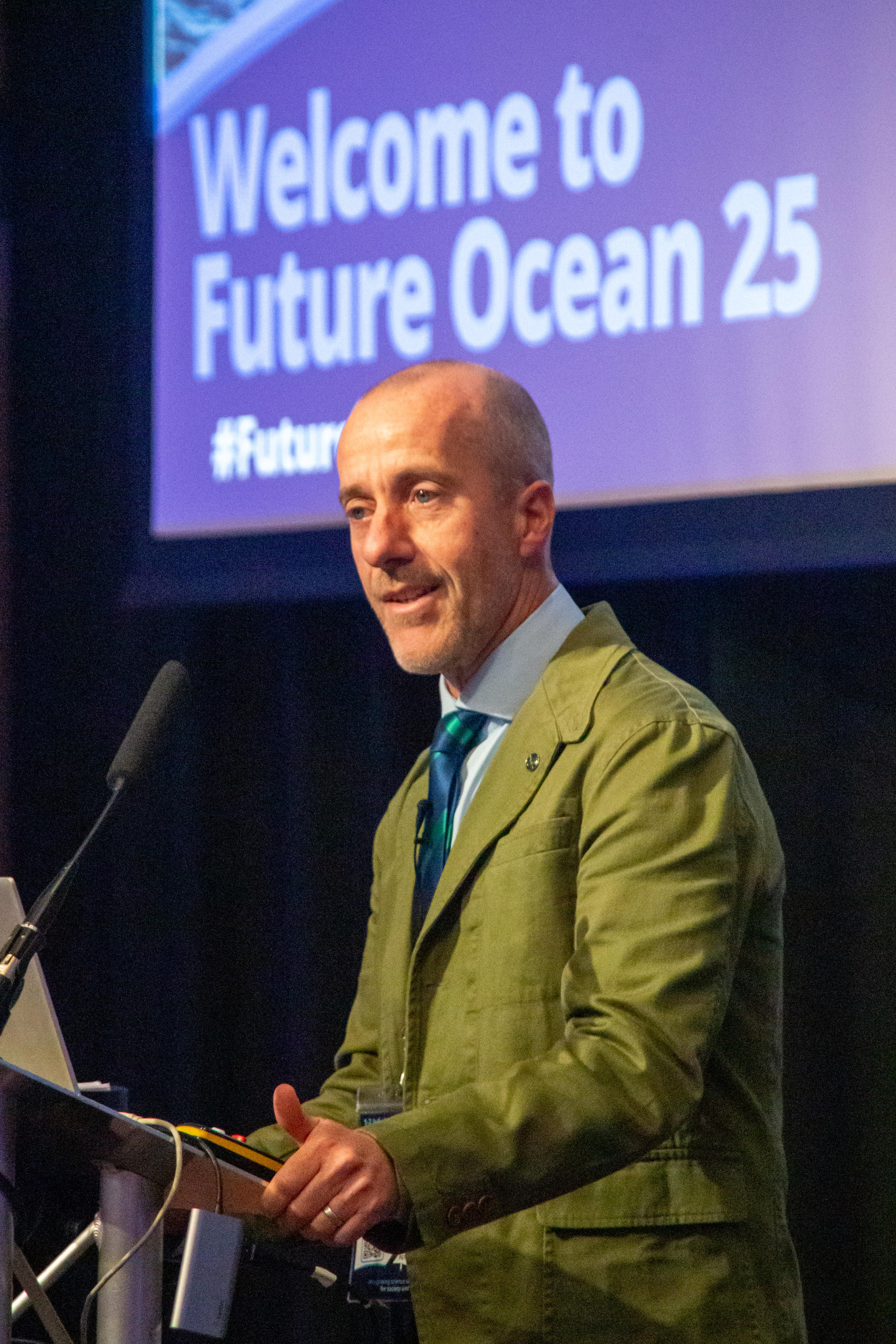 The Chief Scientist at Cefas, Grant Stentiford, giving a speech in front of a large screen which says 'Welcome to Future Ocean 25"