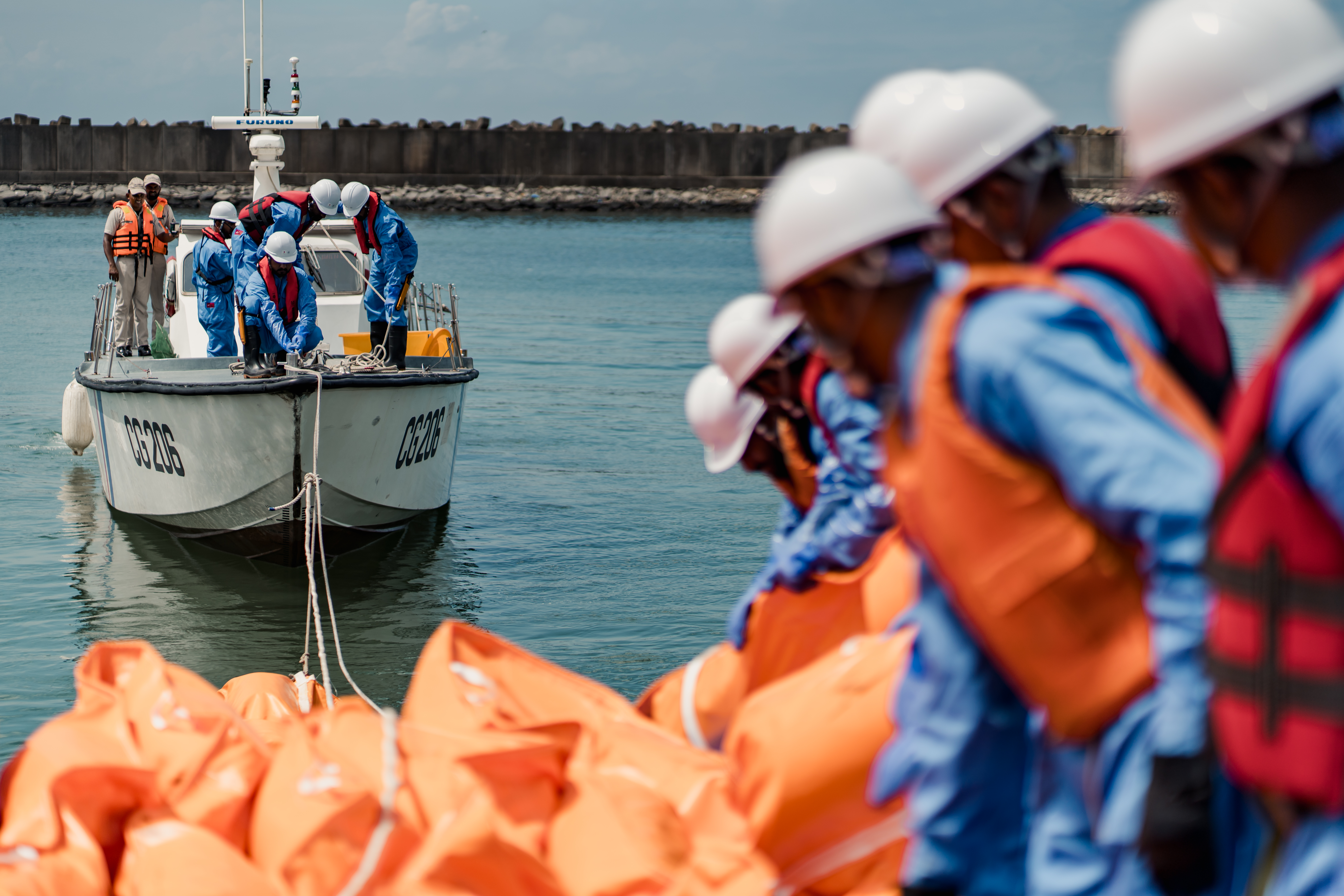 an orange float is being deployed by a boat pulling it with rope, and a group of people standing on land in PPE lowering it into the water.